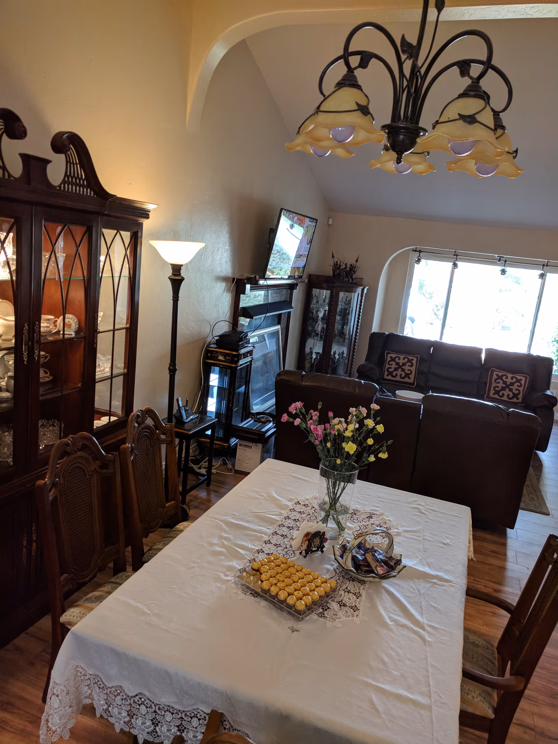 Interior view of a senior living facility showing a dining area with a table covered by a white lace tablecloth, decorated with a vase of flowers and trays of snacks. Behind the dining area is a living room with brown leather sofas, a TV mounted on the wall, a floor lamp, and wooden display cabinets with glass doors containing dishes and decorative items.