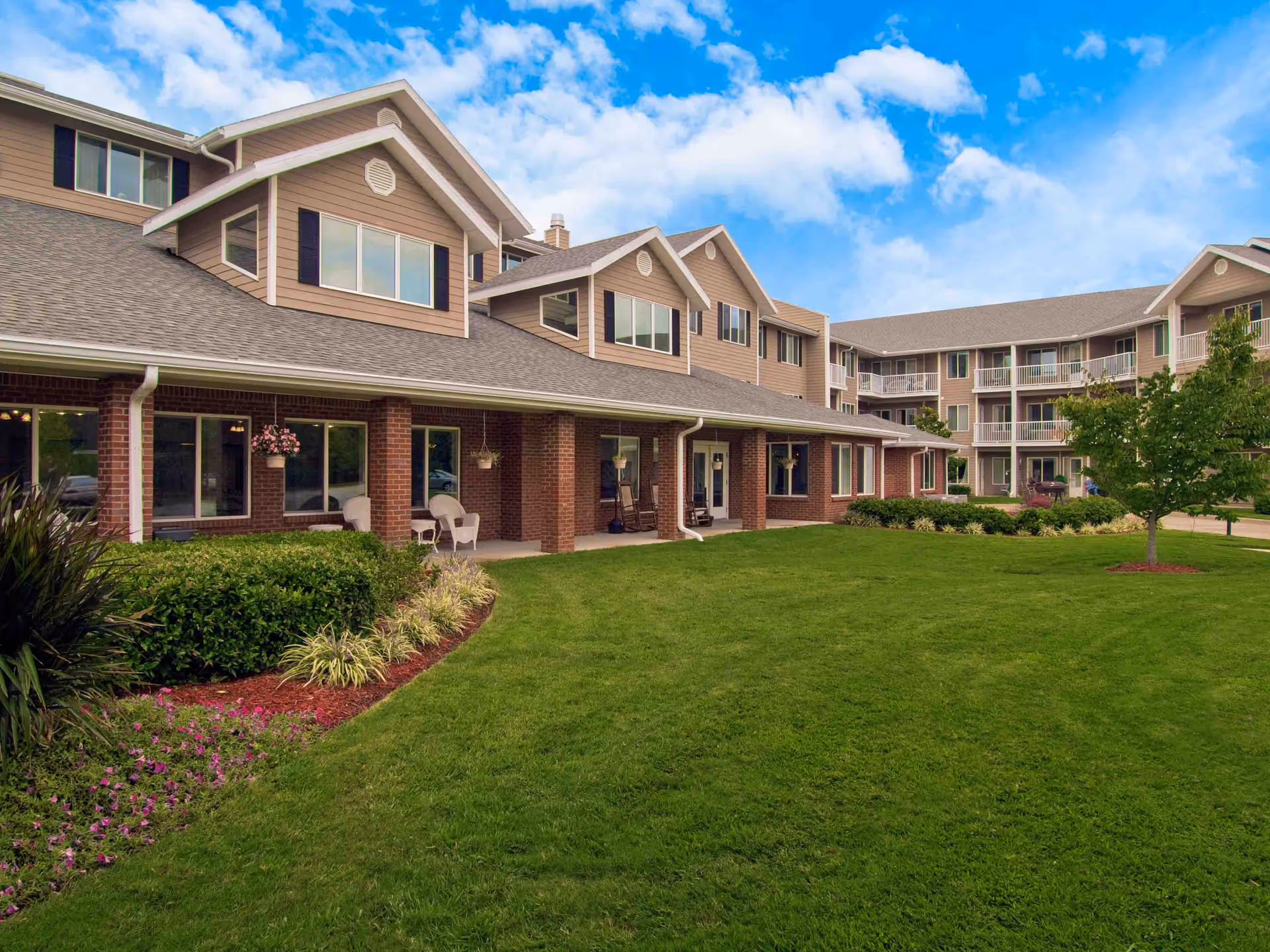 Exterior view of Holiday Pinegate senior living facility showing a well-maintained lawn, landscaped garden beds with flowers and shrubs, and a multi-story building with balconies and large windows under a partly cloudy blue sky.