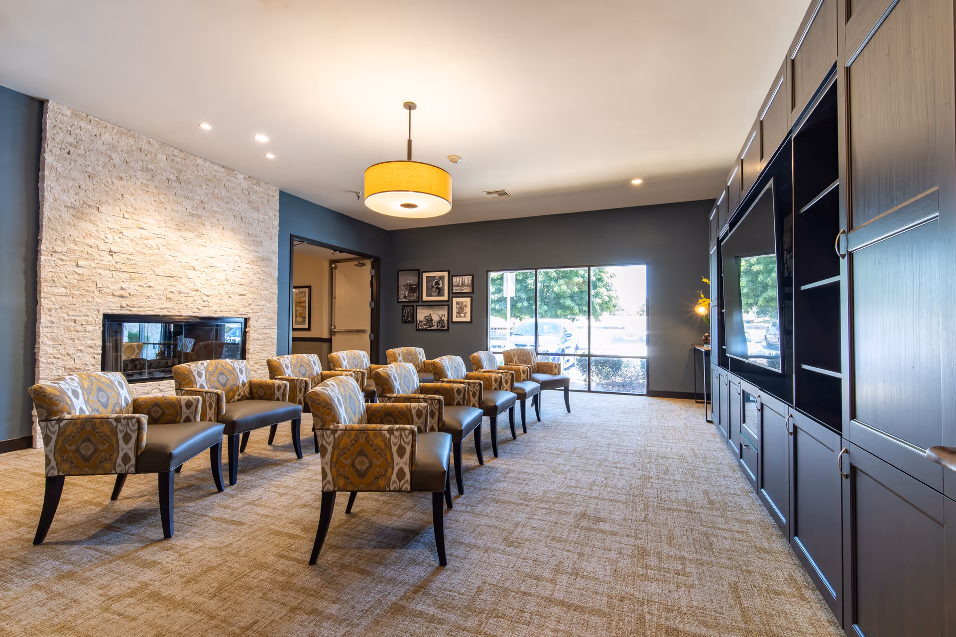 A spacious, well-lit common area with multiple patterned armchairs arranged in rows facing a large built-in entertainment center with a TV. The room features a modern stone fireplace on one side, a large window letting in natural light, and a ceiling light fixture.
