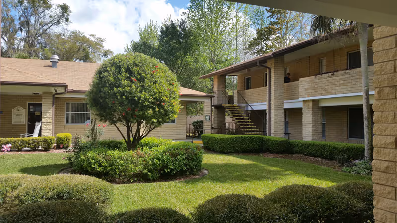 View of a courtyard garden area within a senior living facility, featuring a well-maintained lawn, trimmed bushes, a small tree with red flowers, and beige brick buildings surrounding the garden. There is an outdoor staircase leading to the second floor of one building, and the sky is partly cloudy.