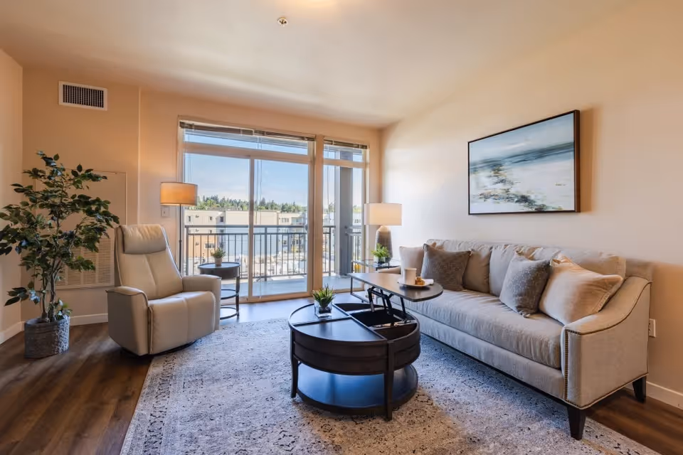 A bright living room with a beige sofa adorned with multiple cushions, a round dark wood coffee table with a lift-top, a beige recliner chair, two side tables with lamps, a large window and glass door leading to a balcony, and a large framed artwork on the wall.