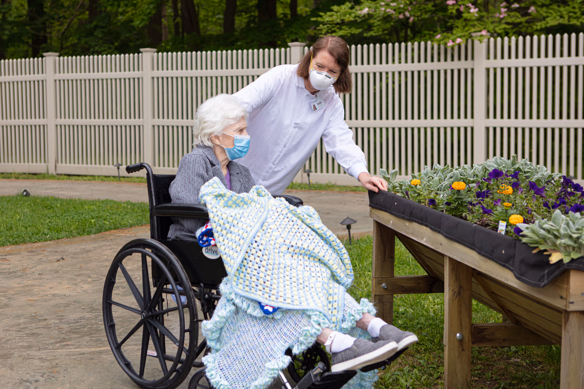 An elderly woman in a wheelchair covered with a blanket is outdoors next to a raised garden bed with flowers. A caregiver wearing a white coat and a face mask is standing beside her, pointing at the flowers. Both are wearing face masks and are near a white fence with green trees in the background.