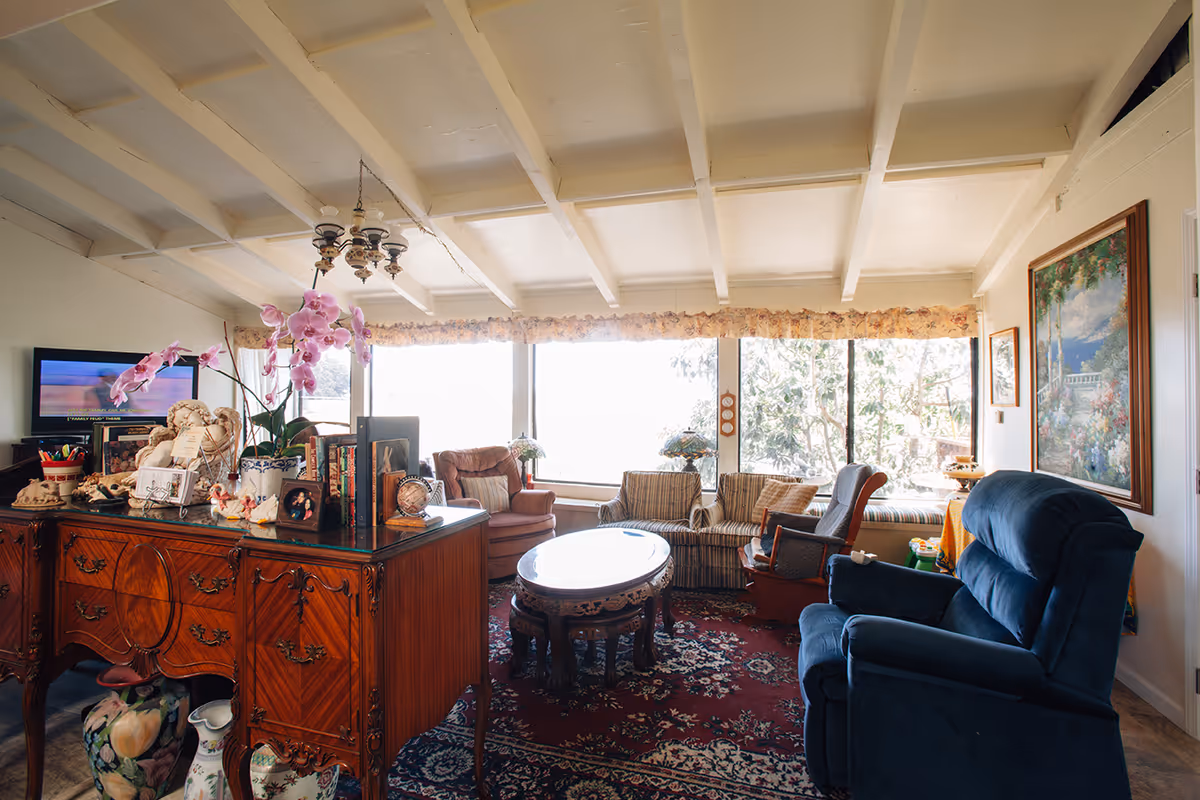 Sunlit, cozy living room with large windows, upholstered chairs, an ornate wooden sideboard topped with flowers and decor, and a central round coffee table.