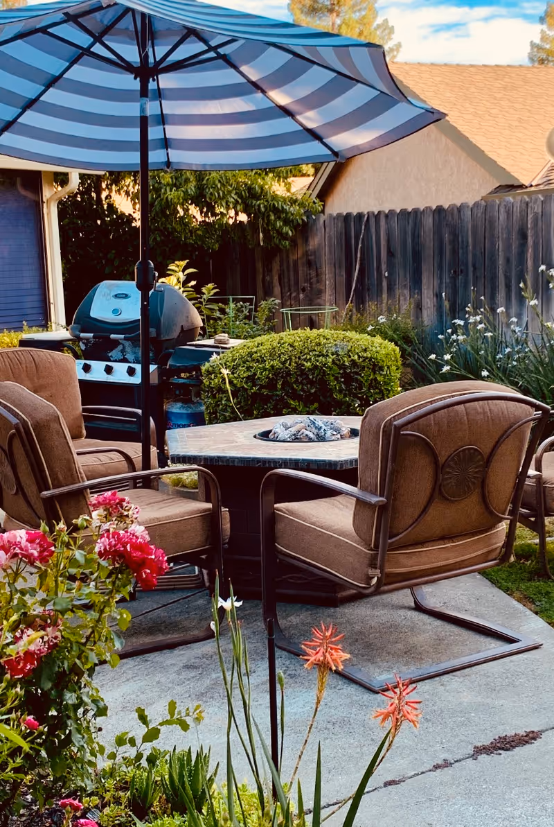 Backyard patio with a round fire-pit table surrounded by cushioned chairs under a striped umbrella, a grill, and flowering plants.