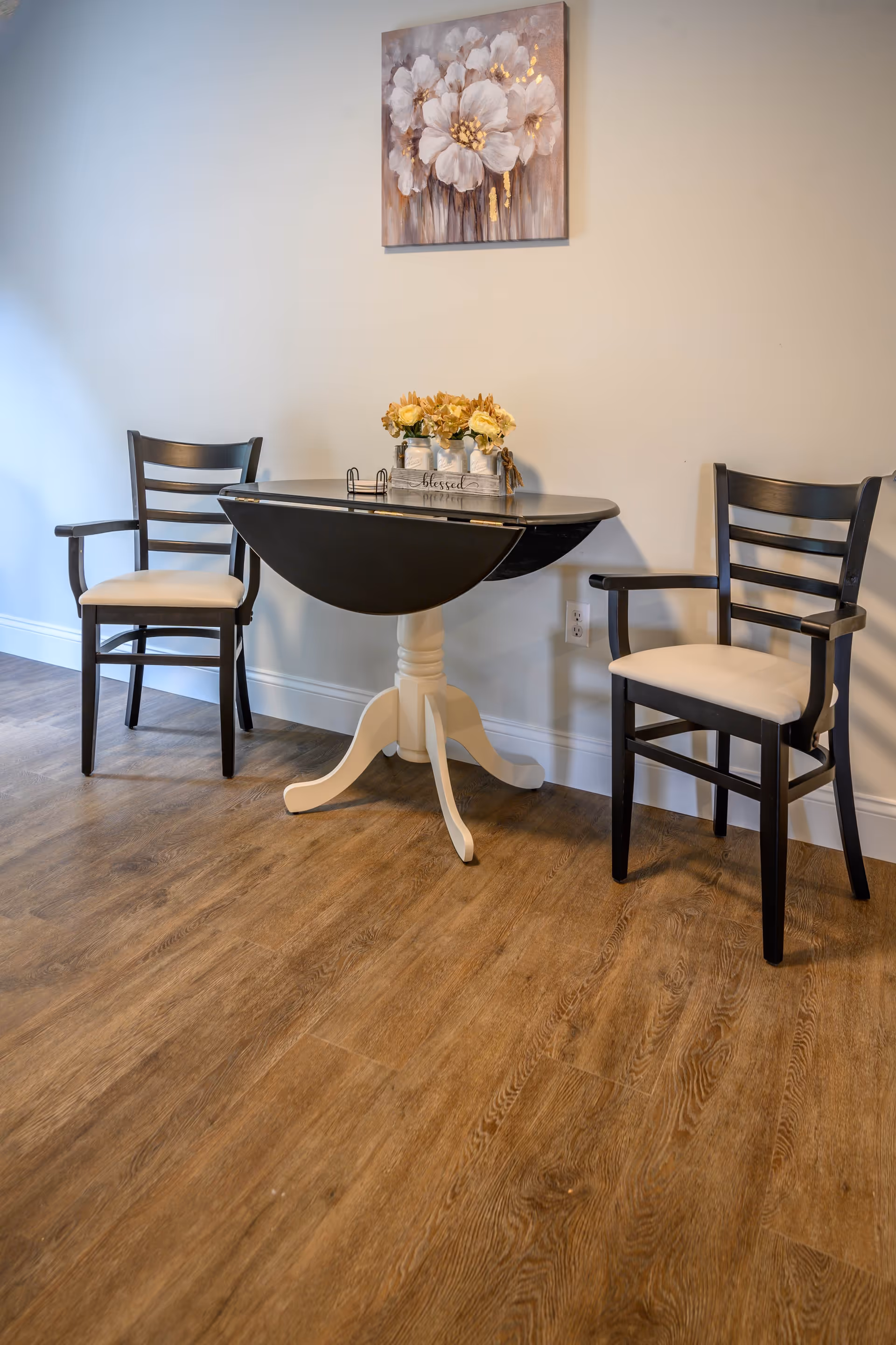 A small dining area with a round black and white drop-leaf table and two black wooden chairs with beige cushions. On the table is a decorative arrangement of flowers in white vases inside a wooden tray with the word 'blessed' written on it. Above the table hangs a painting of large white flowers with gold accents. The floor is wooden and the walls are painted light beige.