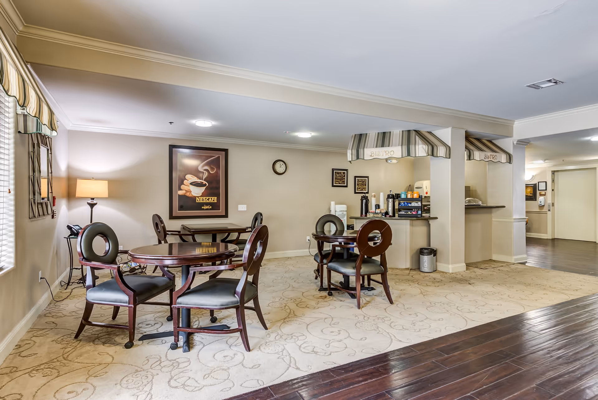 Interior view of a cozy seating area in a senior living facility named Cypress Place, featuring round wooden tables with four chairs each, a coffee station labeled 'Bistro' with striped awnings, a framed coffee-themed artwork on the wall, a floor lamp, and a carpeted floor with a decorative pattern next to dark wooden flooring.