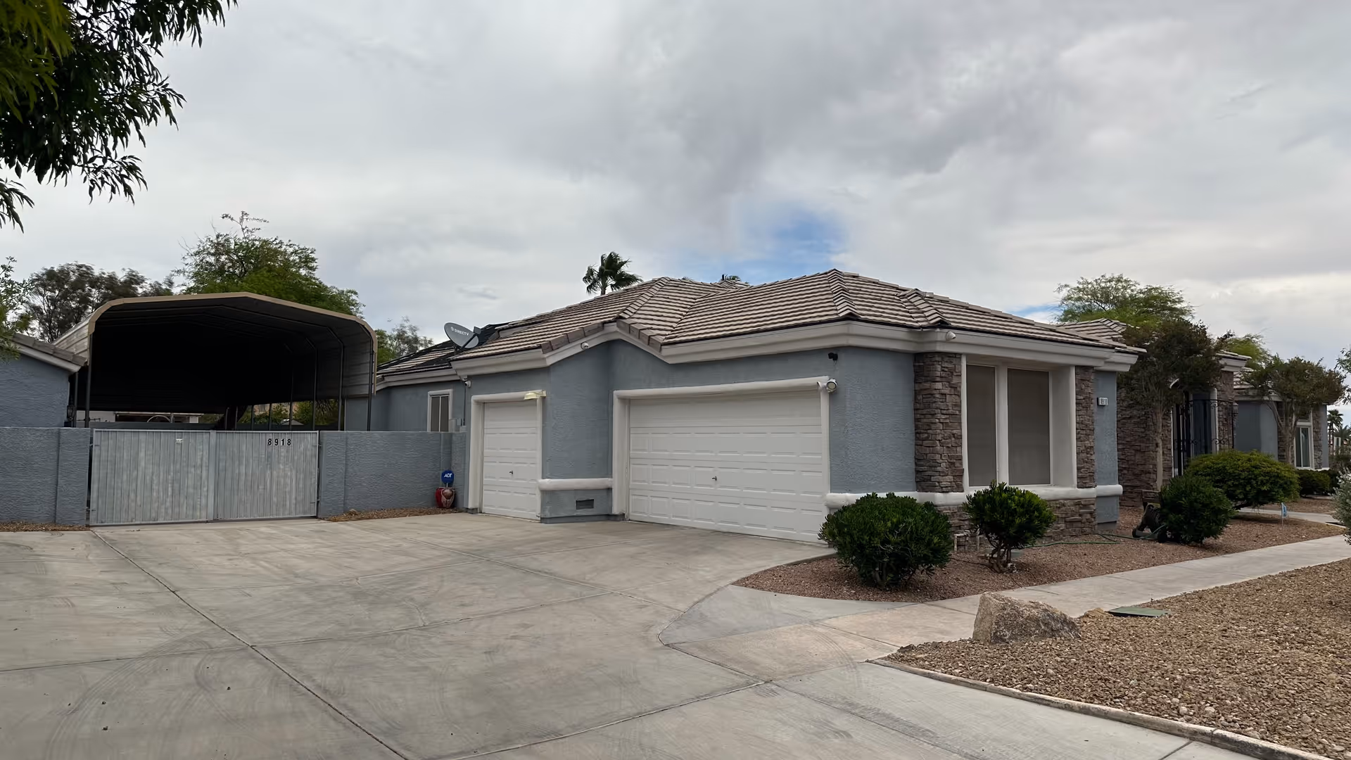 Exterior view of a single-story senior living facility building with a tiled roof, two white garage doors, and stone accents on the walls. There is a concrete driveway and a gated carport on the left side. Small bushes and desert landscaping with gravel surround the building under a cloudy sky.