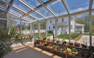Sunlit glass-enclosed sunroom with potted plants on a bench overlooking a white house and landscaped yard.