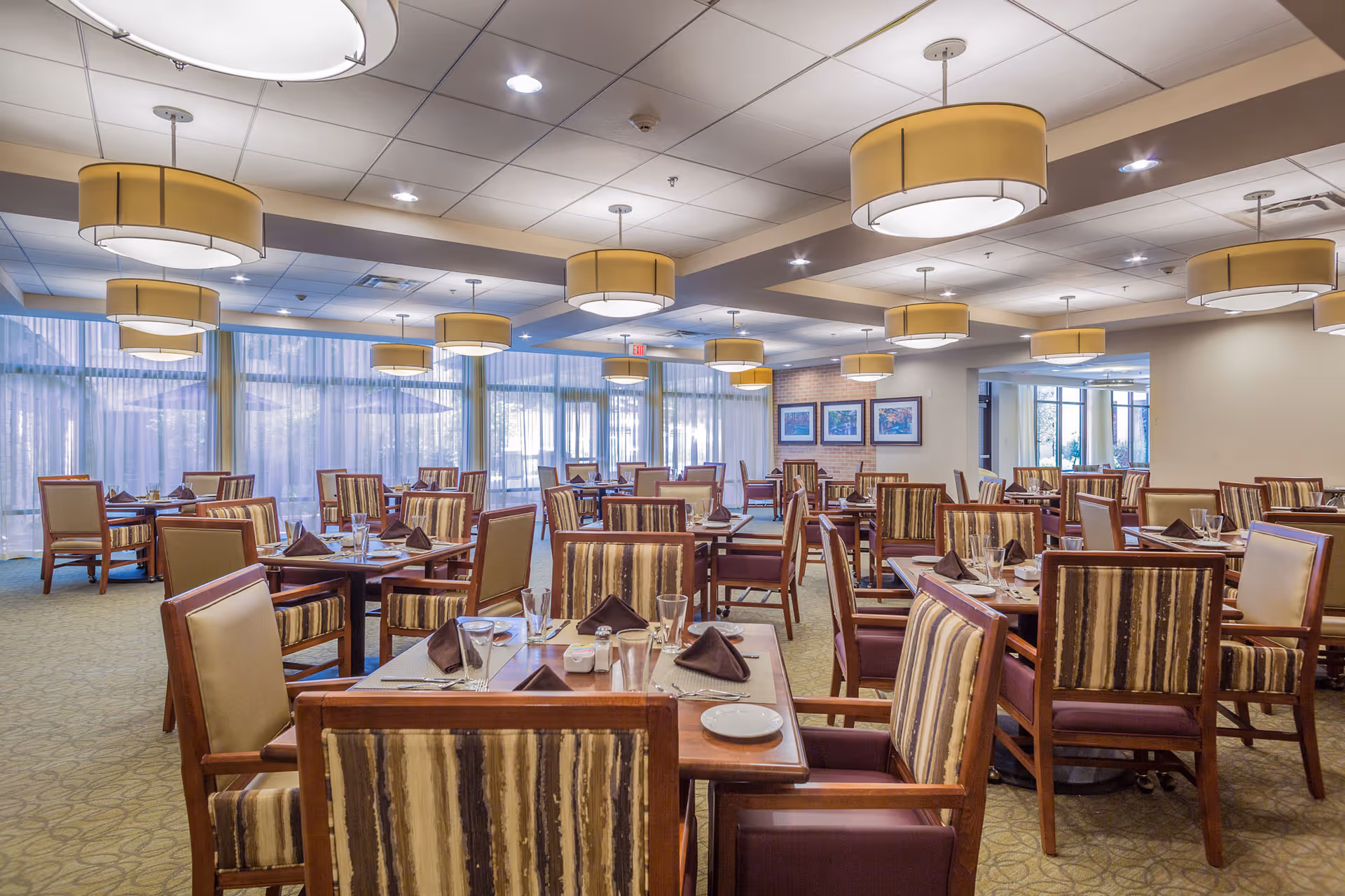 Spacious dining room with multiple set tables and striped upholstered chairs under hanging drum lights.