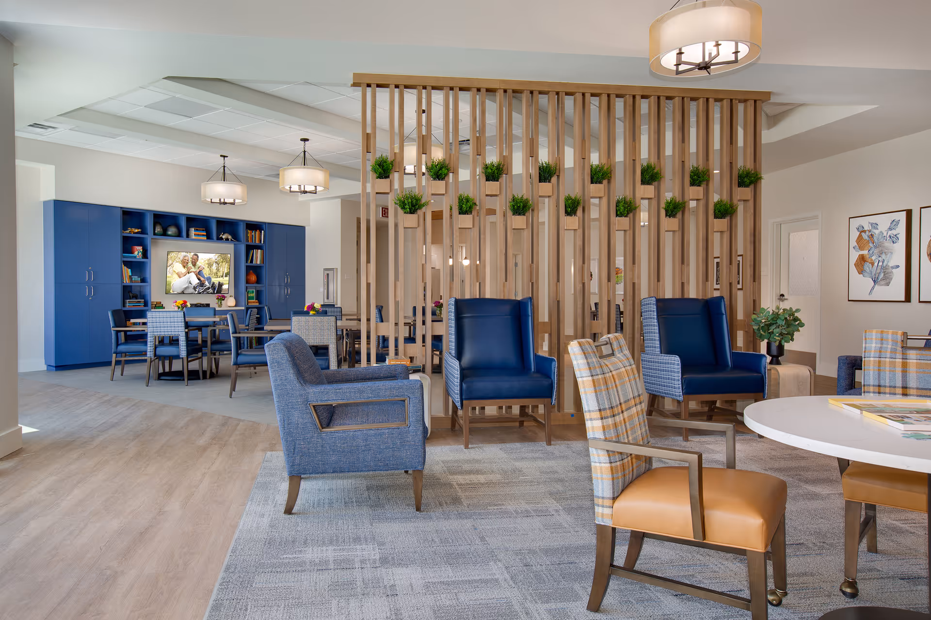 A modern senior living facility common area with a mix of blue and plaid upholstered chairs around a white round table and a wooden slatted room divider adorned with small green plants. In the background, there are more tables and chairs, a blue built-in cabinet with shelves holding books and decorative items, and a TV displaying an image of two people sitting on grass. The room has light wood flooring and neutral walls with framed artwork.