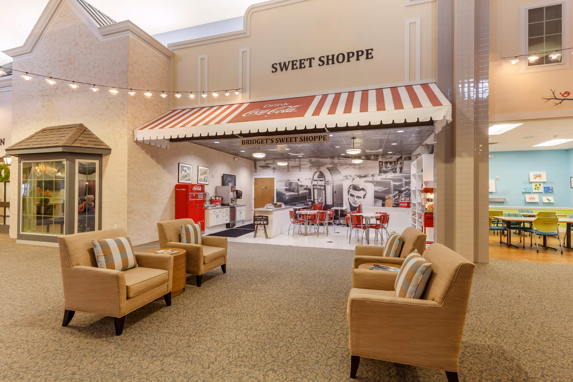Interior view of a senior living facility featuring a cozy seating area with four beige armchairs and a small table in the center. In the background, there is a retro-style sweet shoppe with a red and white striped awning labeled 'SWEET SHOPPE' and 'BRIDGET'S SWEET SHOPPE'. The shop has red chairs, a vintage Coca-Cola refrigerator, and a black and white mural of a classic diner scene with a jukebox and a portrait of a man. String lights hang above the seating area, and a dining area with tables and chairs is visible to the right.