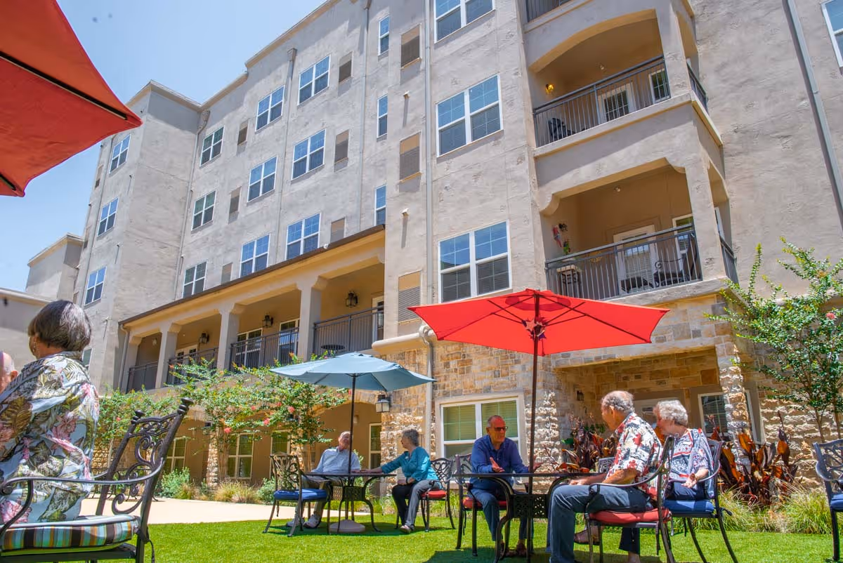 Seniors sitting at tables with umbrellas in a courtyard outside a multi-story apartment building.