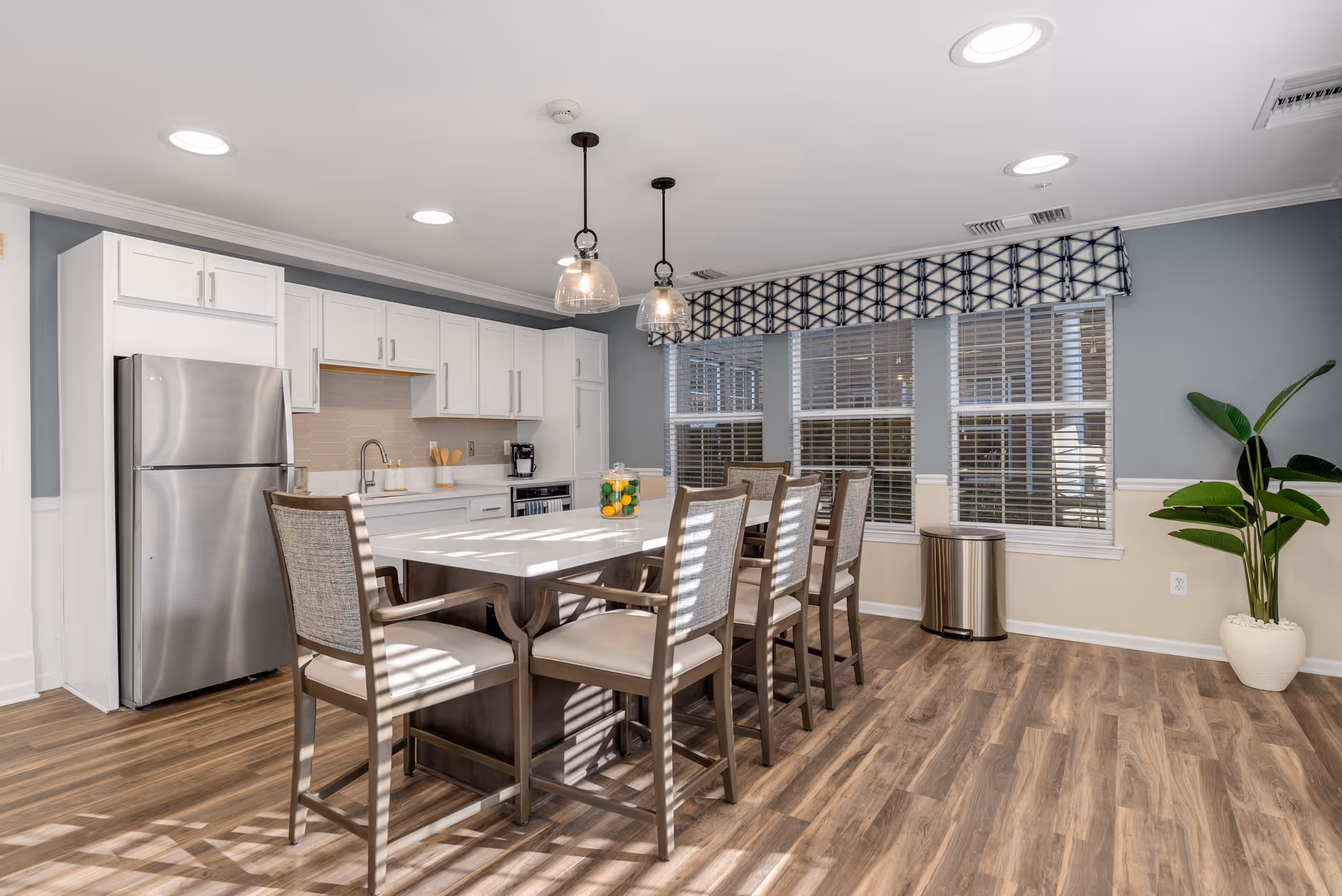A bright and modern kitchen and dining area with white cabinets, stainless steel refrigerator, and a large island with seating for six. The room features wood flooring, three large windows with blinds and a patterned valance, two pendant lights hanging over the island, and a potted plant in the corner.