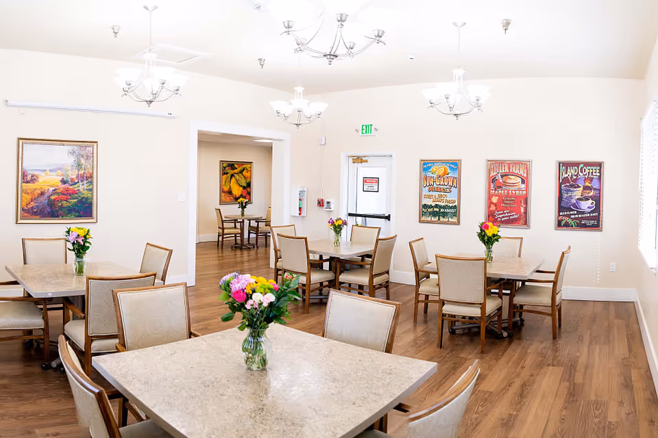 A bright dining room in a senior living facility with multiple square tables, each surrounded by four chairs. Each table has a vase with colorful flowers. The room has wooden flooring, cream-colored walls, and several framed paintings and posters on the walls. There are multiple ceiling light fixtures and an exit door at the back.