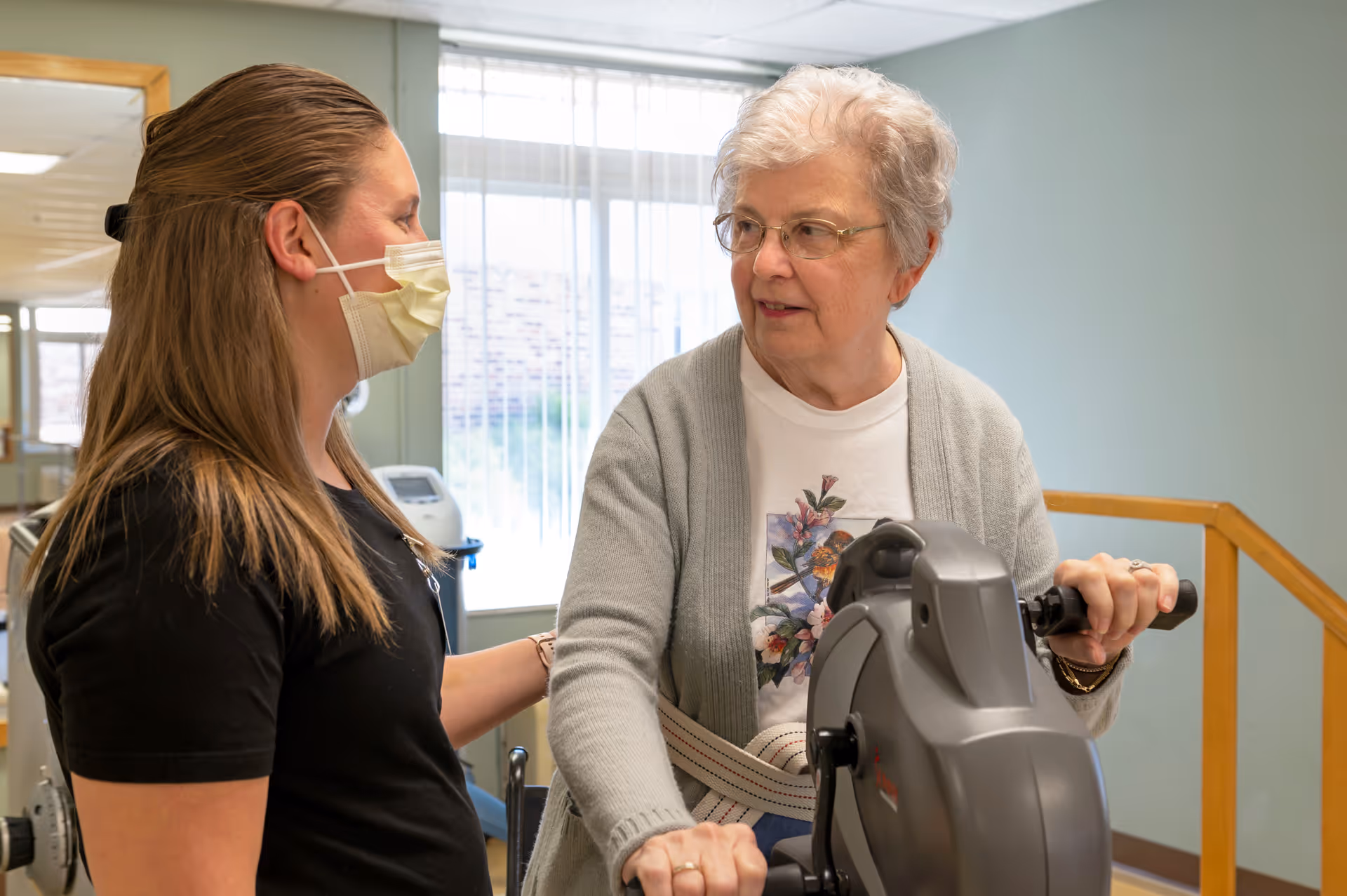 An elderly woman using a stationary exercise bike with assistance from a healthcare worker wearing a face mask in a rehabilitation center room with light green walls and a window with vertical blinds.