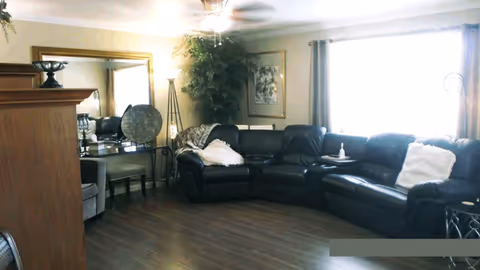 A cozy living room with a large black leather sectional sofa adorned with white pillows. The room features wooden flooring, a tall floor lamp, a large mirror on the wall, framed artwork, and a potted plant in the corner. Natural light streams in through a window with curtains.