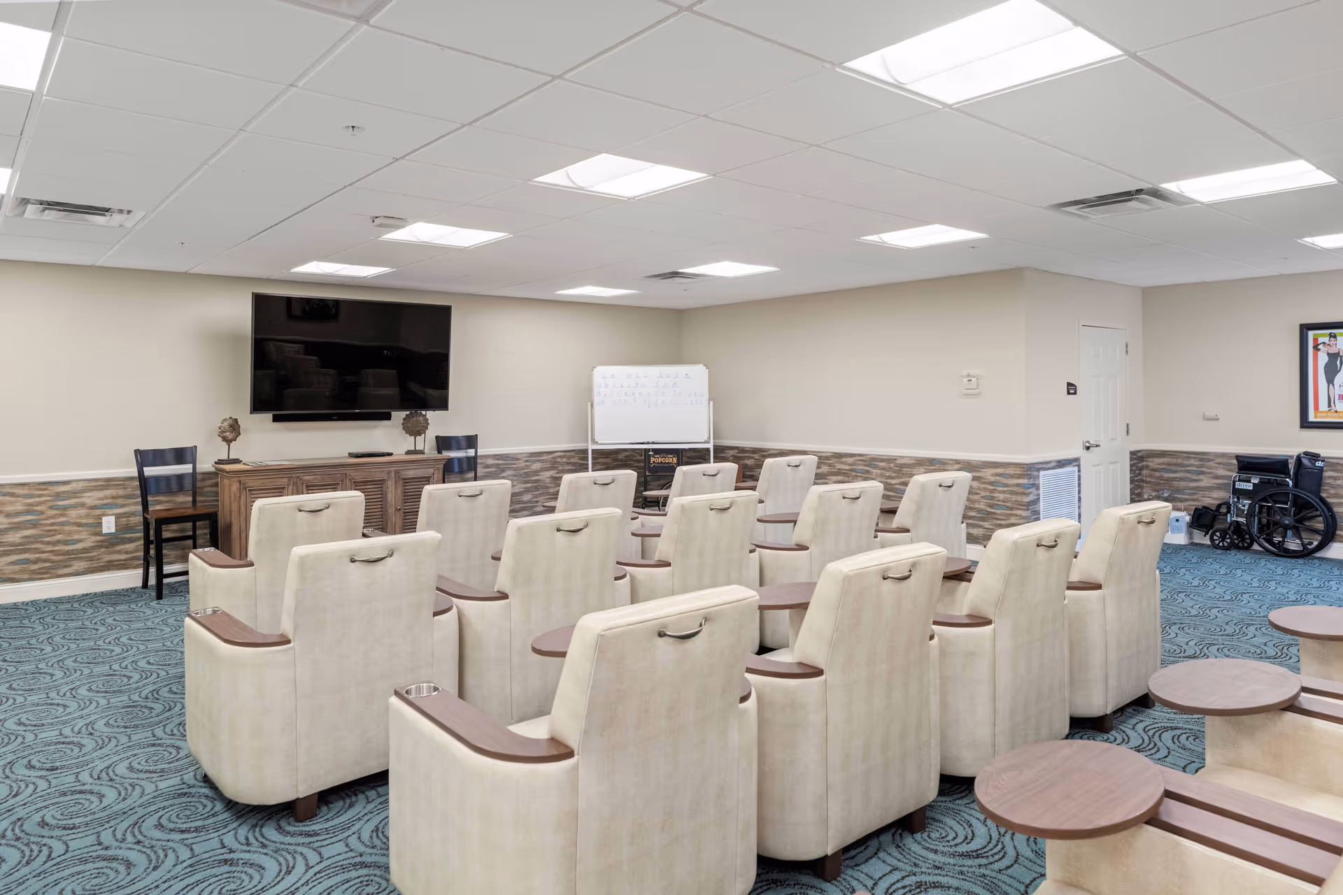 A senior living facility common room with multiple beige armchairs arranged in rows facing a large flat-screen TV mounted on the wall. There is a wooden cabinet beneath the TV, a whiteboard on a stand in the corner, and a wheelchair near the wall. The room has patterned blue carpet and neutral-colored walls with a chair rail and wainscoting.