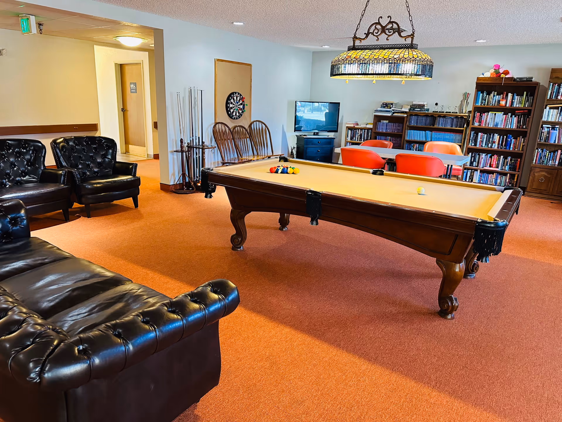 A cozy recreational room with a pool table in the center, surrounded by black leather chairs and a sofa. There is a dartboard on the wall, a TV on a stand, bookshelves filled with books, and a table with orange chairs. The room has warm orange carpeting and a decorative hanging lamp above the pool table.