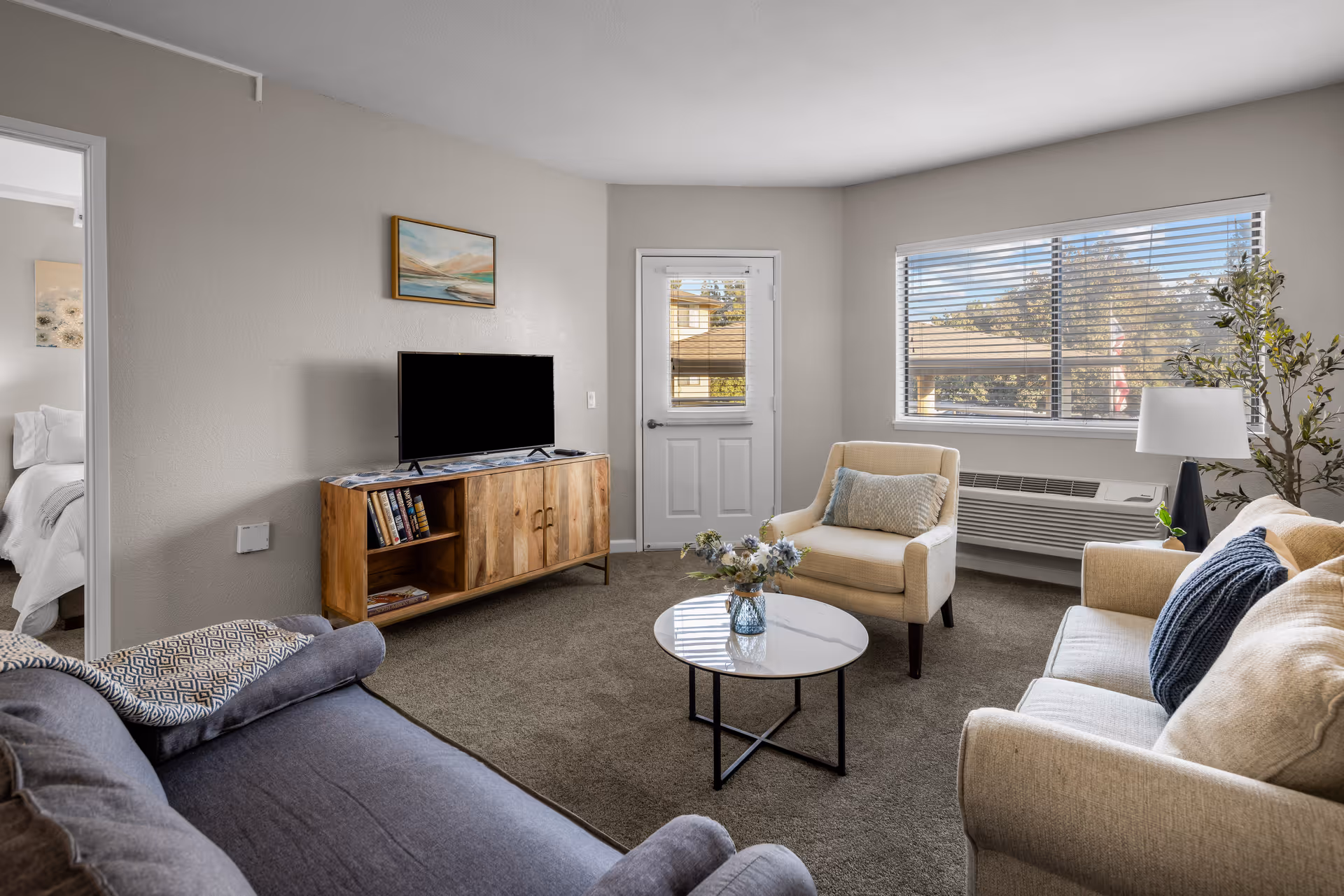 A cozy living room in a senior living community featuring a gray sofa, a beige armchair, a beige loveseat, a round white coffee table with a flower vase, a wooden TV stand with a flat-screen TV, a large window with blinds, a door with a window, and a wall painting above the TV stand.