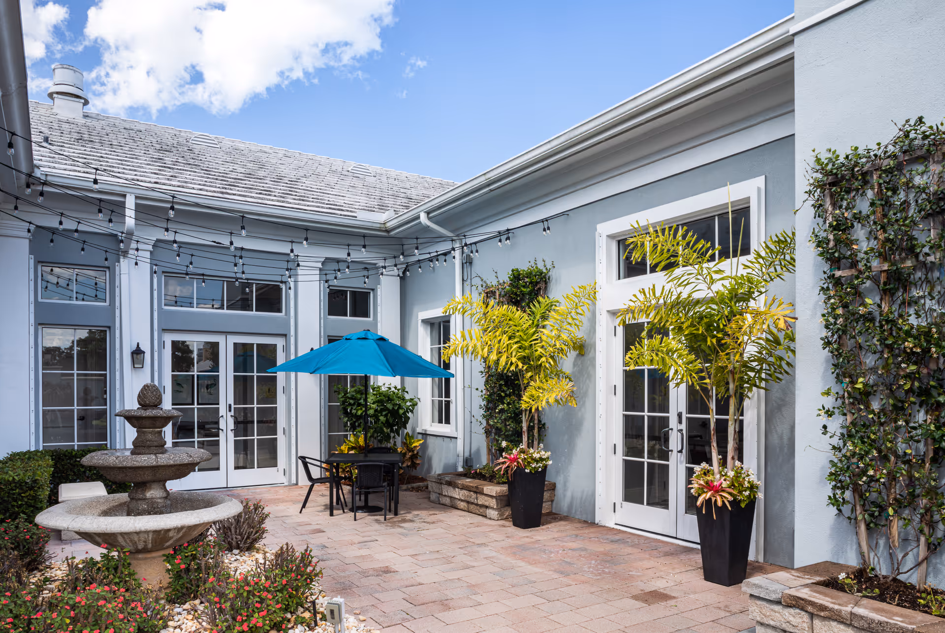 Sunlit courtyard with a stone fountain, a patio table under a blue umbrella, potted plants, and French doors on a light-blue building.