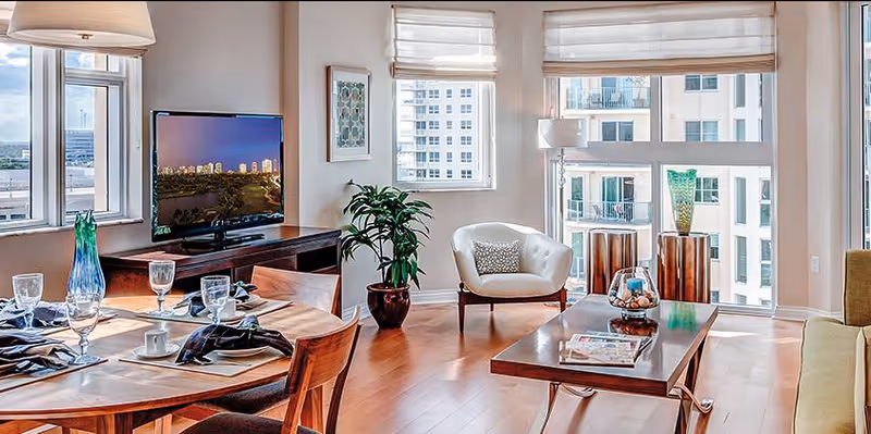 Bright open living and dining area with hardwood floors, a set dining table, TV console, armchair, coffee table and large windows overlooking neighboring buildings.