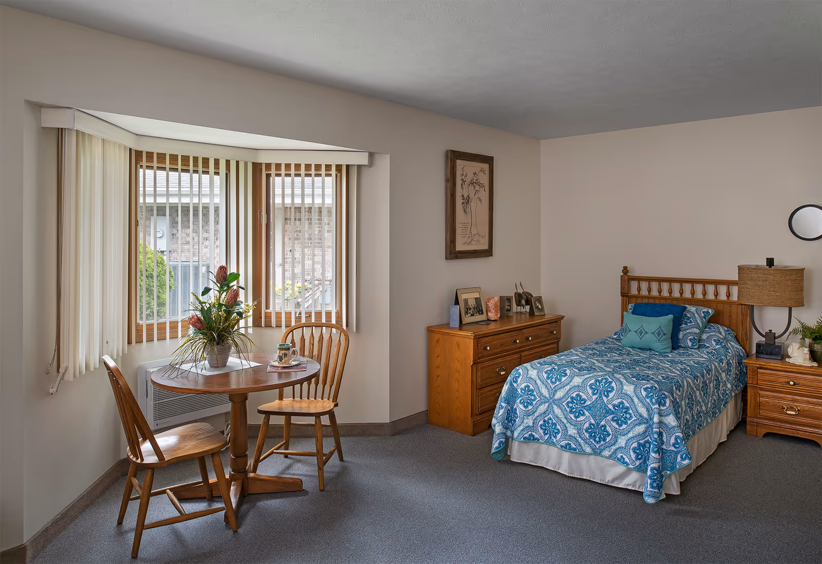 A tidy single bedroom featuring a twin bed with blue patterned bedding, wooden nightstands and dresser, and a small round table with two chairs by a bay window.