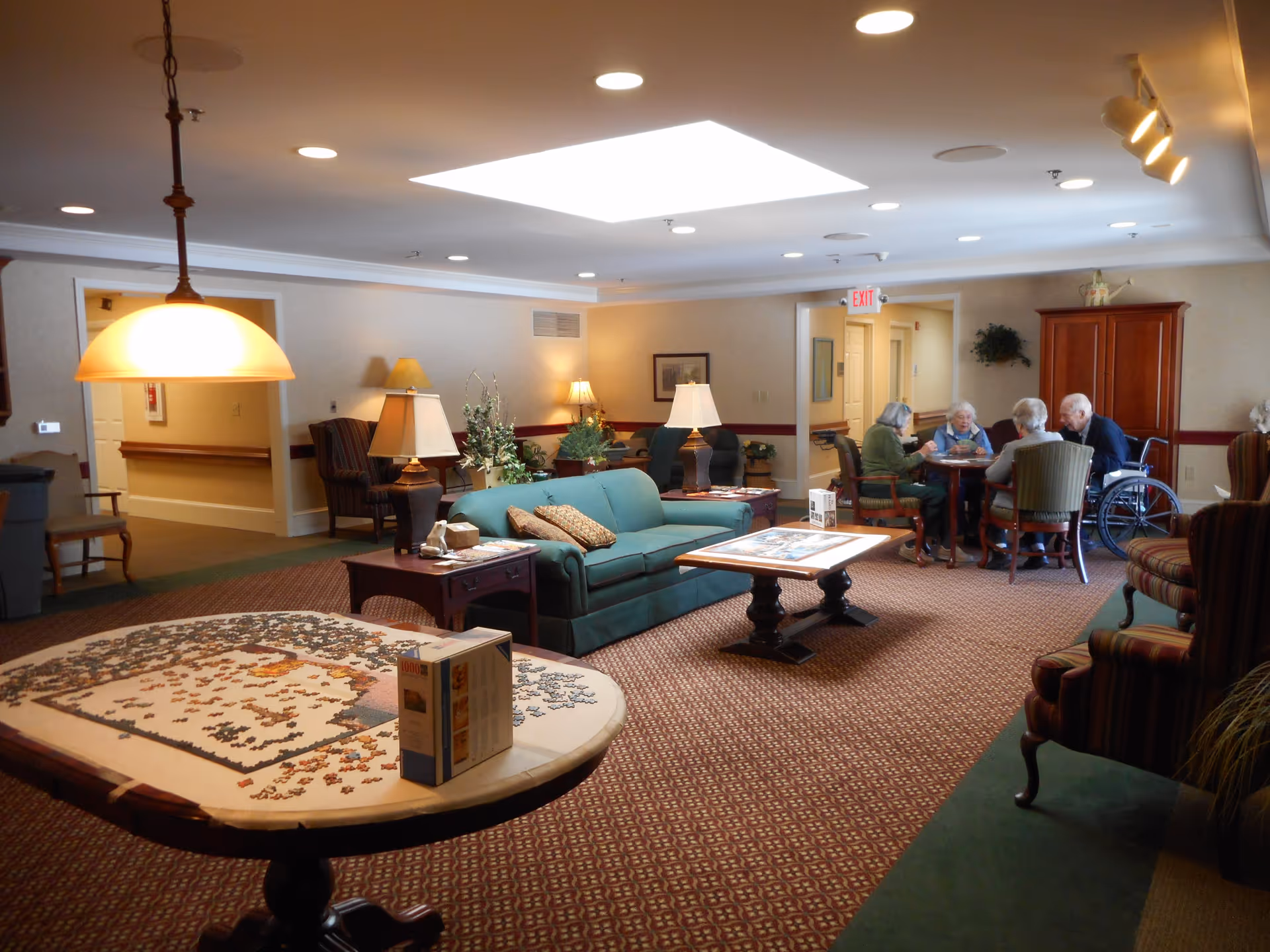 A cozy common area in a senior living facility with a green couch, armchairs, and several lamps. A round table in the foreground has a partially completed jigsaw puzzle on it. In the background, four elderly people are seated around a table, engaged in conversation. The room is warmly lit with ceiling lights and a skylight.