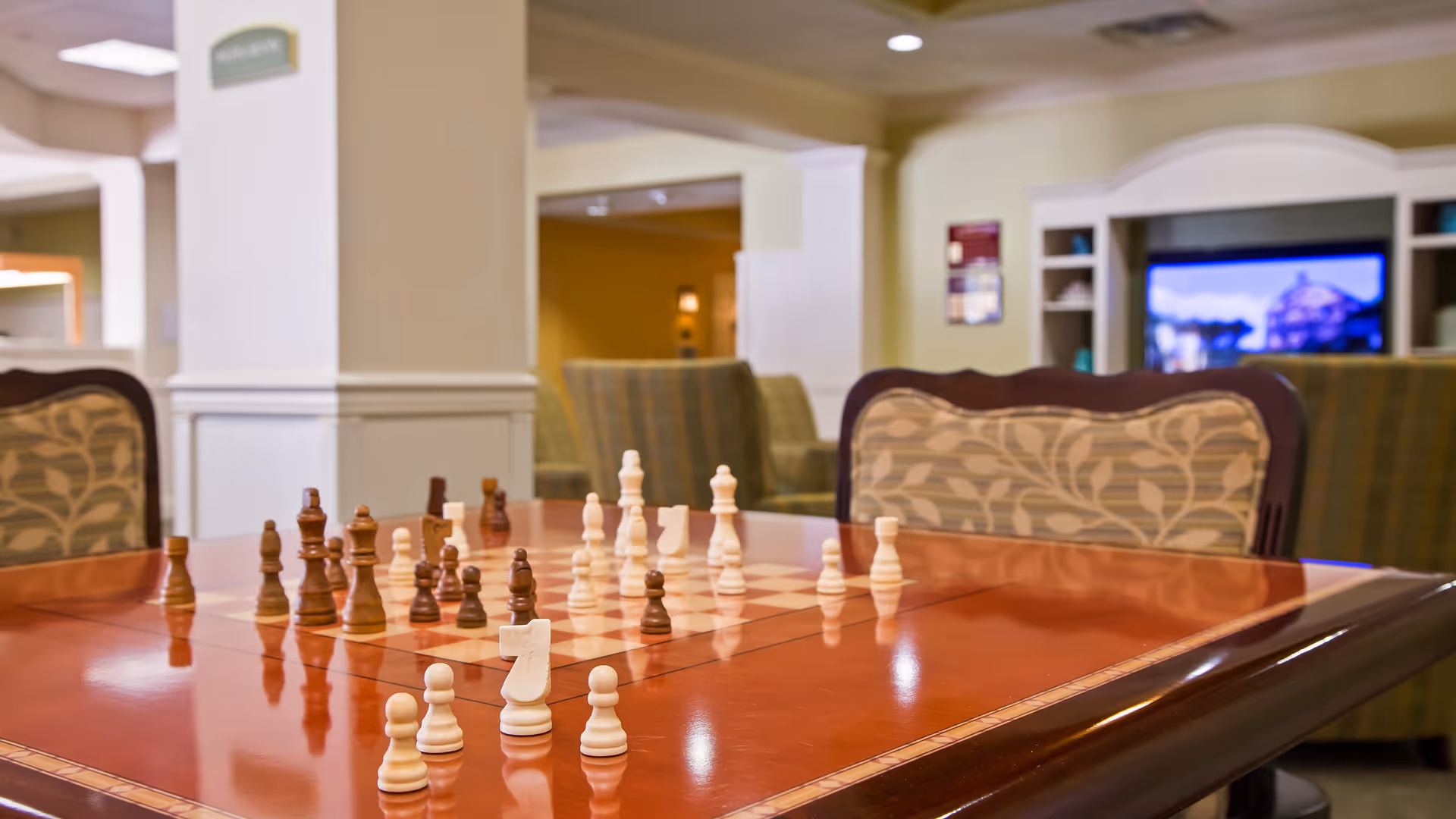 A polished wooden table with a built-in chessboard and chess pieces set up for a game in a cozy living room area with patterned chairs and a TV in the background.