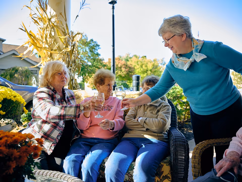 Four elderly women sitting outdoors on wicker furniture, three seated and one standing, raising glasses in a toast. They are surrounded by autumn decorations including yellow flowers and dried cornstalks, with a house and trees visible in the background under a clear sky.