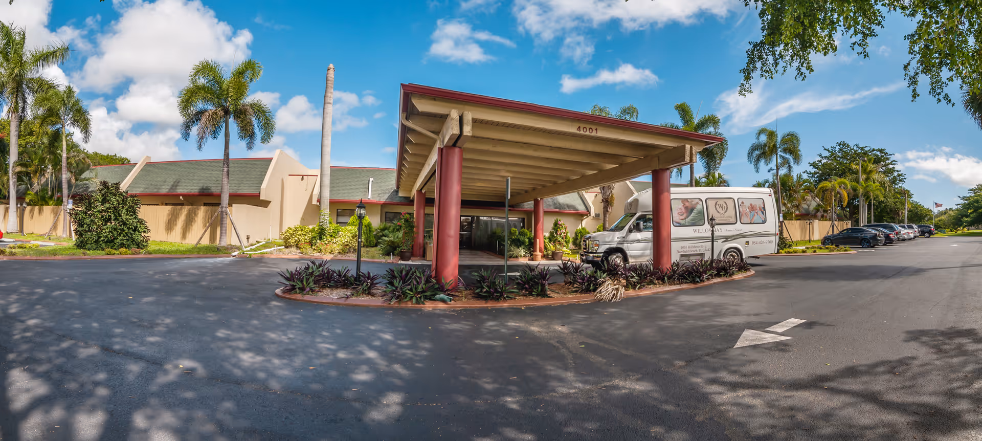 Front entrance of Willow Bay Senior Resort with a covered porte-cochere, a parked shuttle van, palm trees, and cars in the driveway under a blue sky.