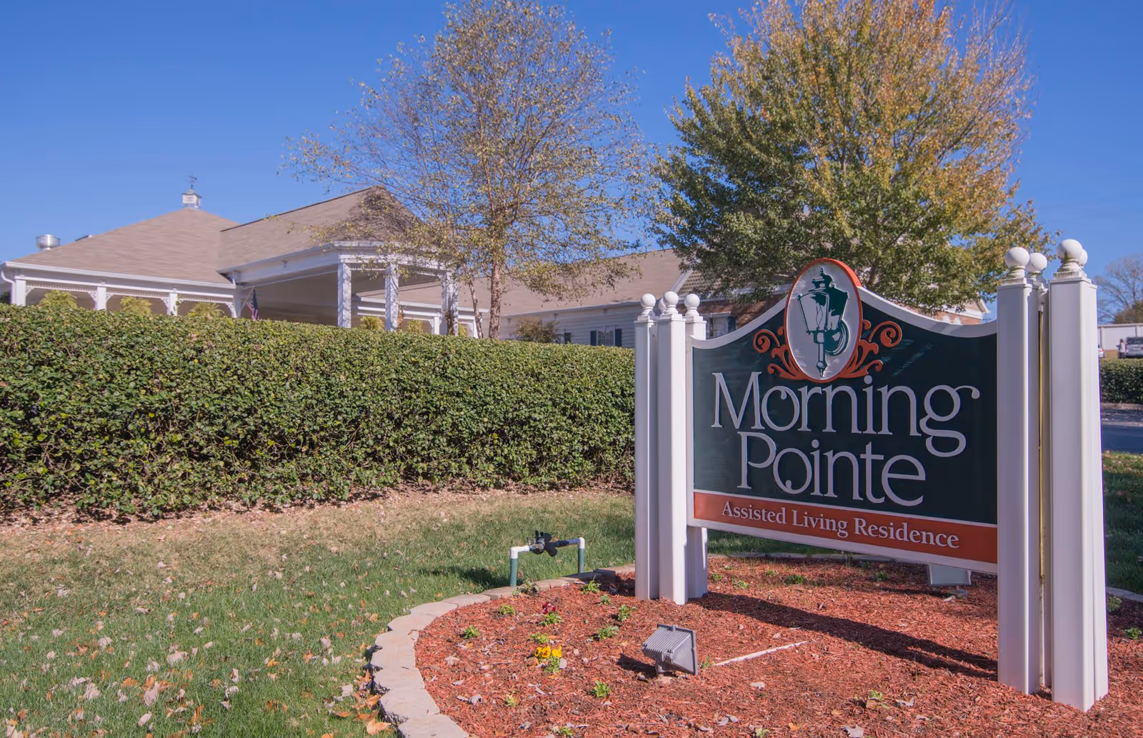 Outdoor view of Morning Pointe Assisted Living Residence sign with a building and trees in the background under a clear blue sky.