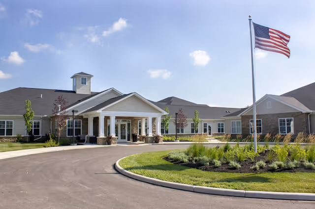 Entrance of a single-story senior living facility with a covered porte-cochère, circular driveway, landscaped center island, and an American flag.