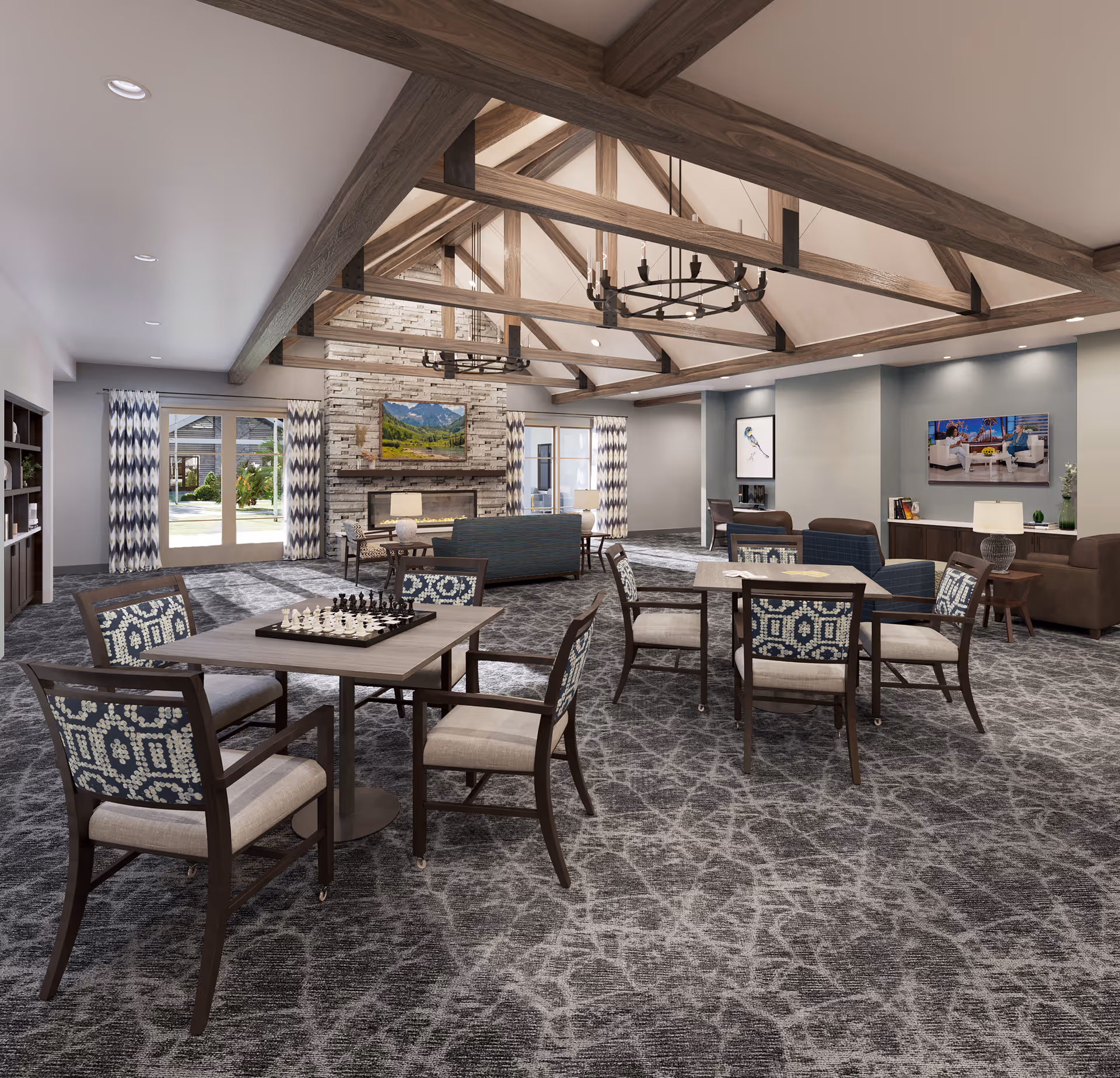 A spacious and well-lit living room area with high vaulted ceilings featuring exposed wooden beams and two large chandeliers. The room has a patterned carpet floor and several seating areas including tables with chairs, one with a chessboard set up. There is a stone fireplace with a mounted TV above it, flanked by large windows with patterned curtains. Additional seating with sofas and armchairs faces another TV mounted on the wall. The room has a cozy and inviting atmosphere with modern decor.