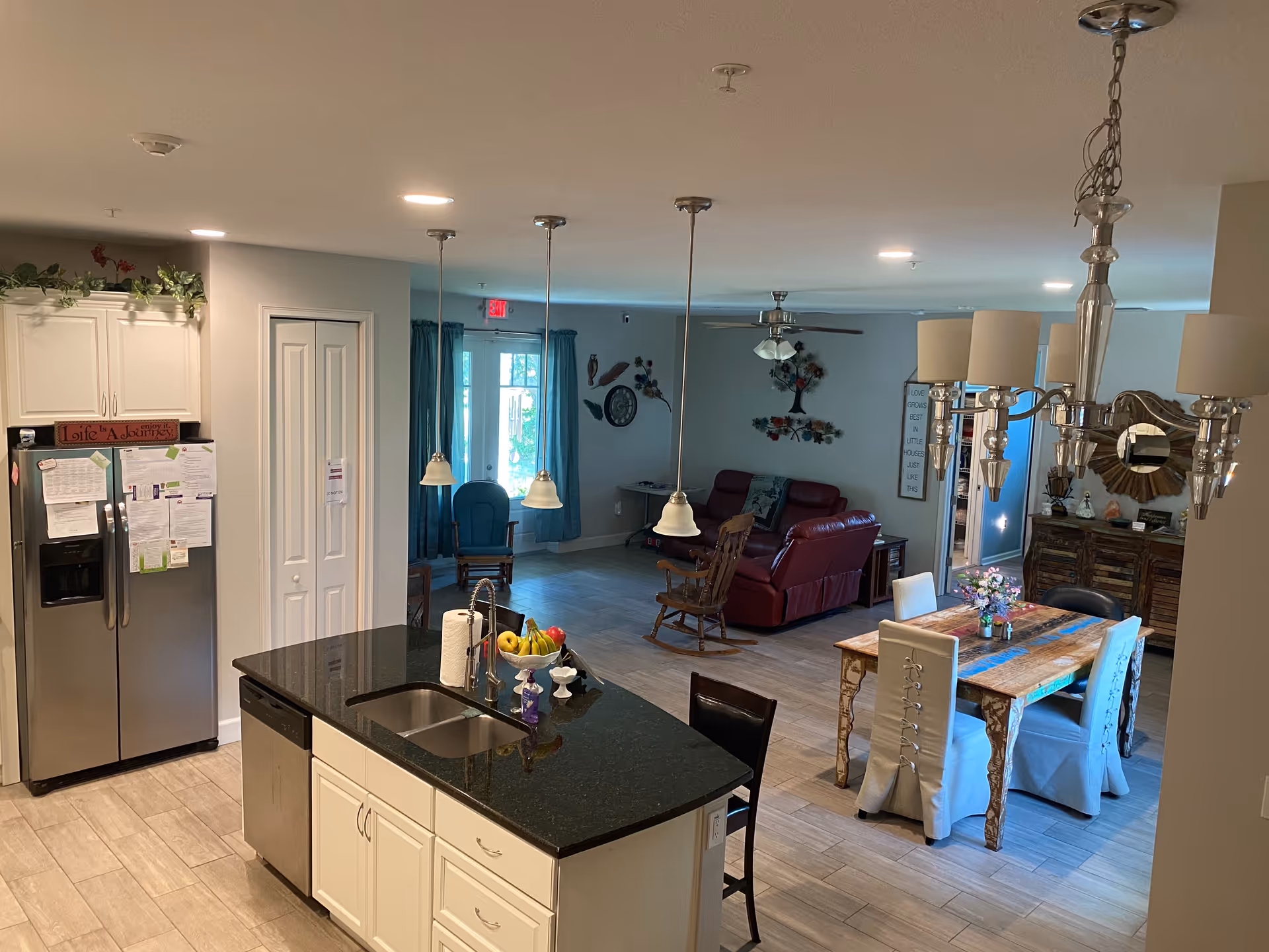 Interior view of a combined kitchen, dining, and living area in an assisted living facility. The kitchen features a black granite island with a sink, paper towels, and a fruit bowl. Stainless steel refrigerator and dishwasher are visible. The dining area has a wooden table with four chairs covered in white slipcovers and a chandelier above. The living area includes a red leather sofa, a rocking chair, wall decorations, and a ceiling fan. Large windows with blue curtains allow natural light into the space.