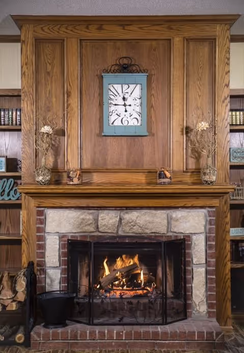 A cozy fireplace with a stone and brick surround, wooden mantel, and a wooden panel above it. On the mantel are decorative items including two glass vases with dried flowers and two small glass candle holders. Above the mantel hangs a large clock with a distressed blue frame. Built-in wooden bookshelves flank the fireplace, holding books and decorative signs.