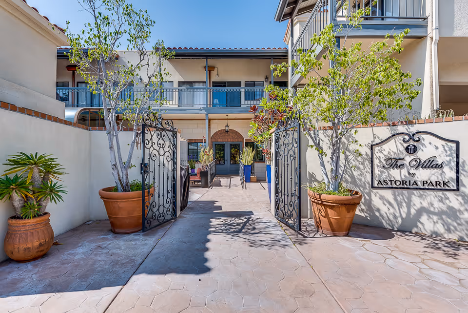 Entrance to The Villas by Astoria Park featuring an open wrought iron gate, potted plants, and a two-story building with balconies in the background under a clear blue sky.