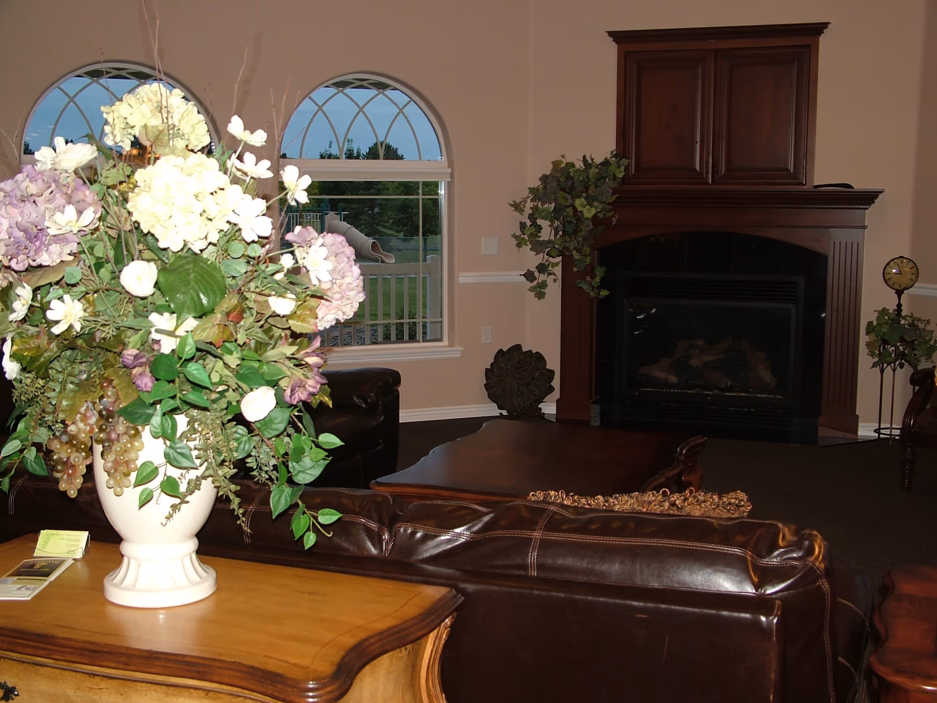 A cozy living room with a large floral arrangement in a white vase on a wooden table in the foreground. Behind it are brown leather sofas and a wooden coffee table. The room features a dark wood fireplace with a plant on top and a decorative clock on a stand to the right. Two arched windows show a view of a playground outside.