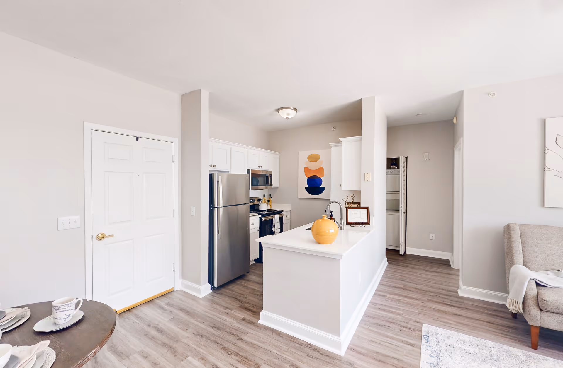 Bright and modern kitchen area with white cabinets, stainless steel refrigerator and microwave, a white countertop island with a yellow decorative vase, and light wood flooring. Adjacent to the kitchen is a small dining table with place settings and part of a living room with a gray armchair and a light-colored rug.