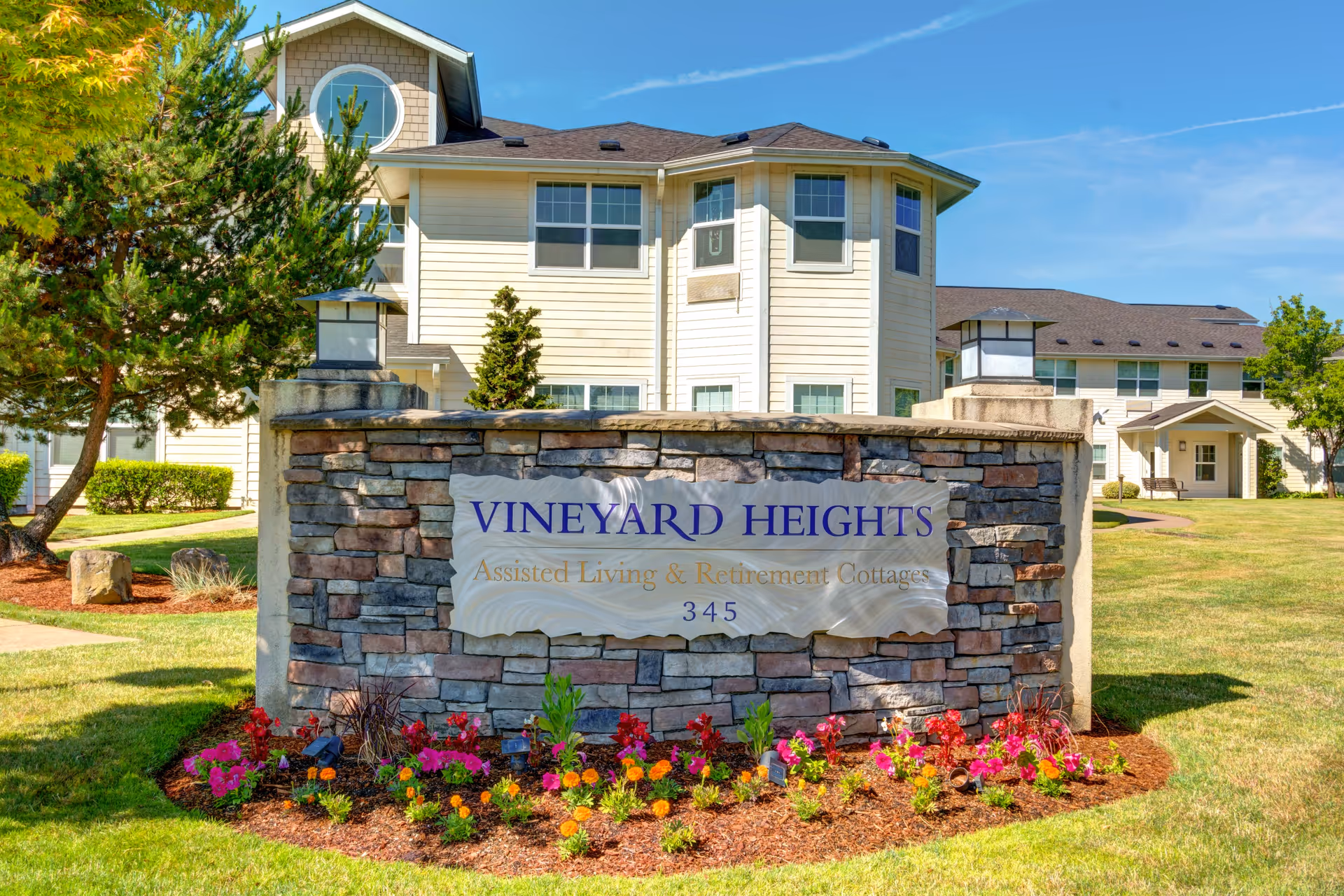 A stone entrance sign reading "VINEYARD HEIGHTS Assisted Living & Retirement Cottages" sits in a flowerbed with the assisted living building and lawn behind it.