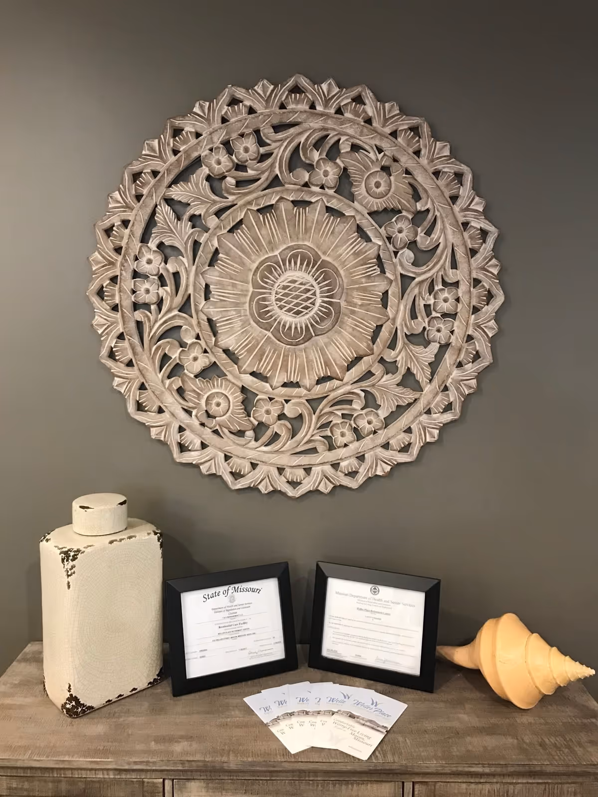A decorative wooden floral wall art hangs above a wooden surface displaying two framed certificates, a large cream-colored ceramic vase with distressed edges, a yellow conch shell decoration, and several business cards for Weller Place Retirement Center.