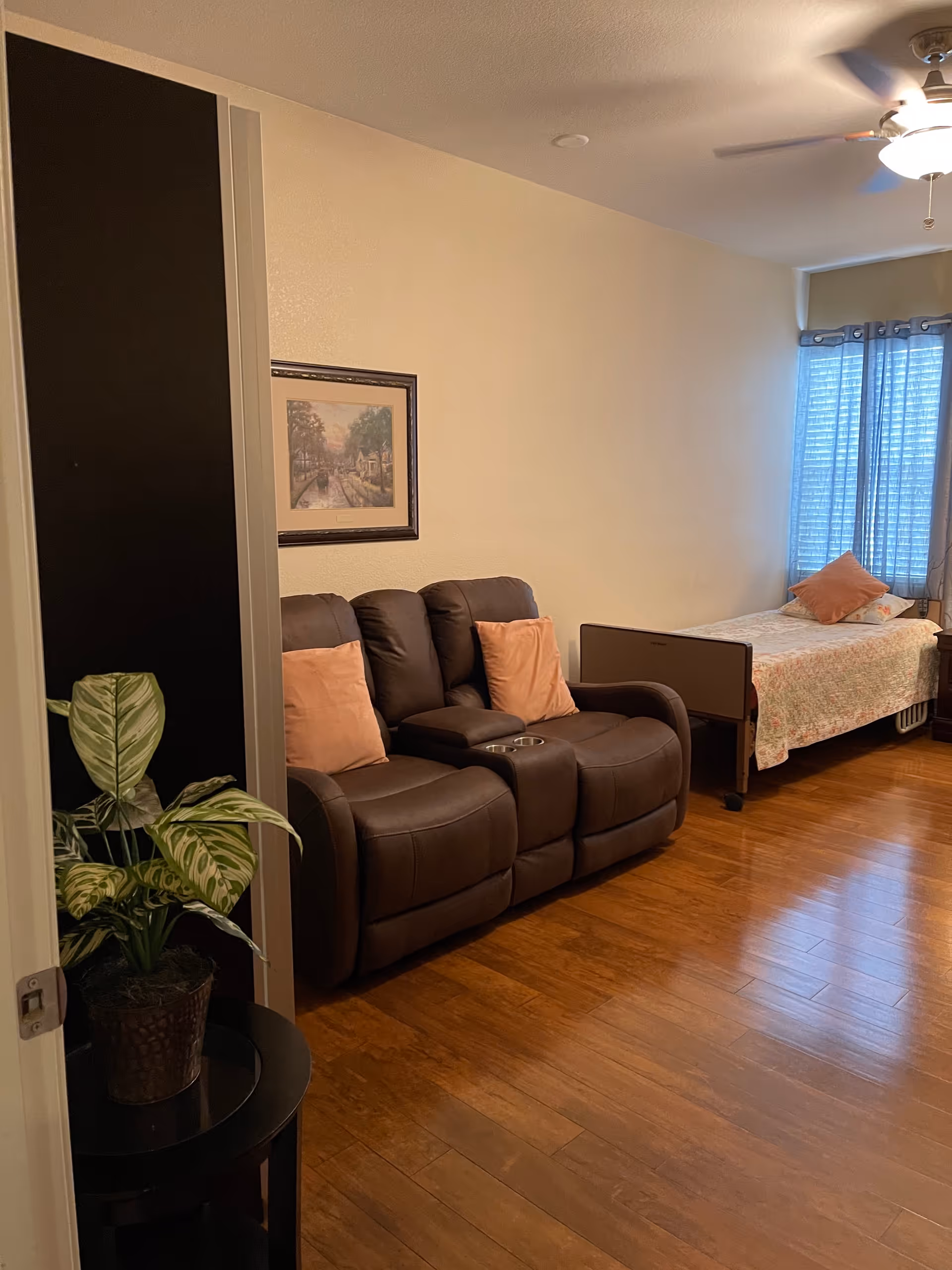 A cozy room with wooden flooring featuring a brown leather loveseat with two peach-colored pillows, a framed painting on the wall above it, a single bed with a floral bedspread and a peach pillow near a window with blue curtains, and a small round table with a potted plant in the foreground.