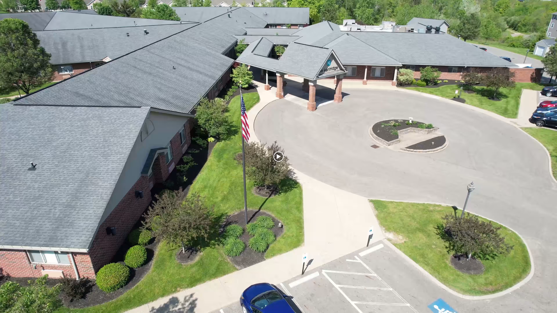 Aerial view of The KentRidge Senior Living front entrance with a circular driveway, flagpole, parking spaces and surrounding landscaping.