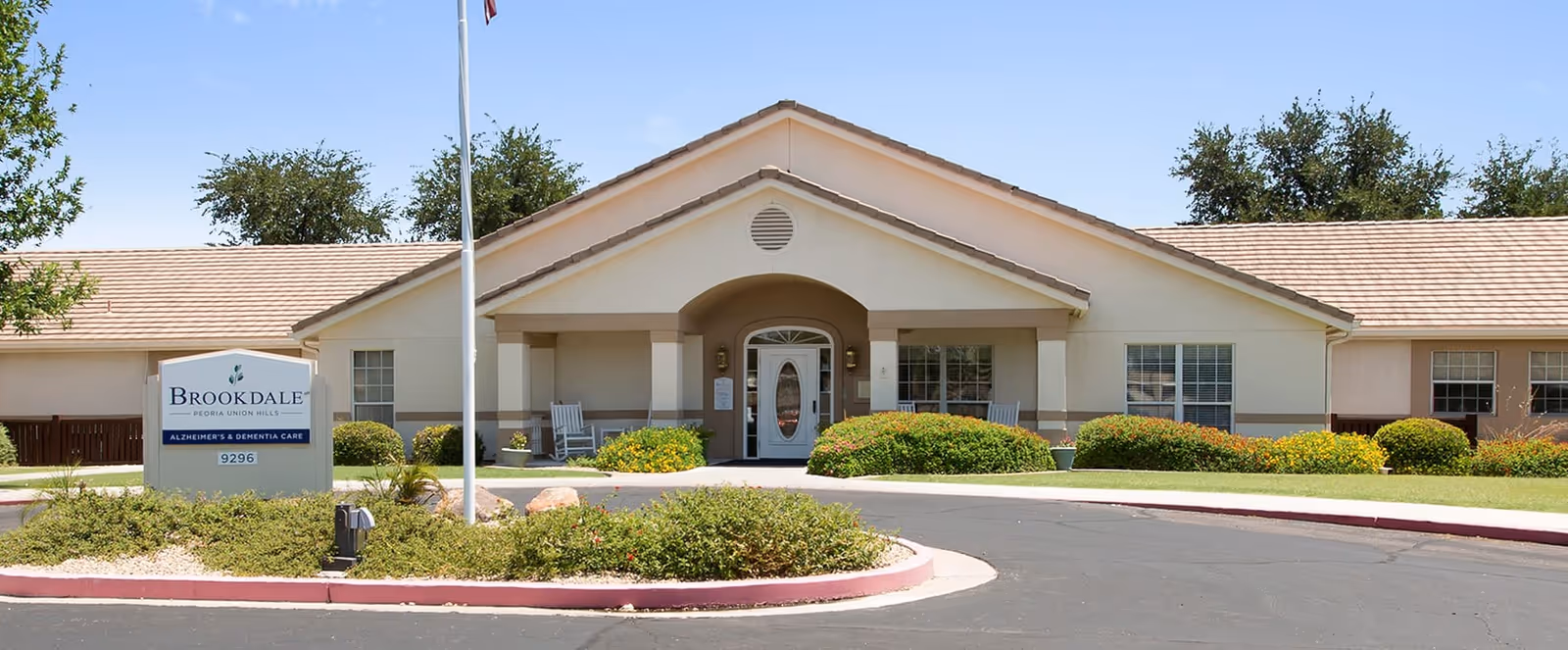 Front exterior view of Brookdale Union Hills facility with a beige building, a covered entrance, a flagpole, and landscaped bushes and greenery in front.
