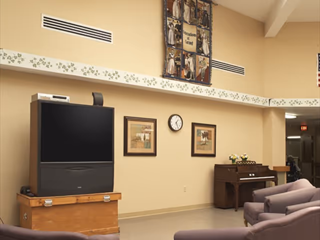 Common living area with a television on a wooden trunk, a piano, seating, a wall clock and framed artwork.