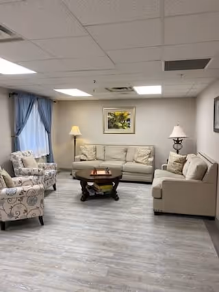 A cozy living room with light-colored sofas and armchairs arranged around a dark wooden coffee table. The room has light wood flooring, a framed picture on the wall, two table lamps, and a window with blue curtains.