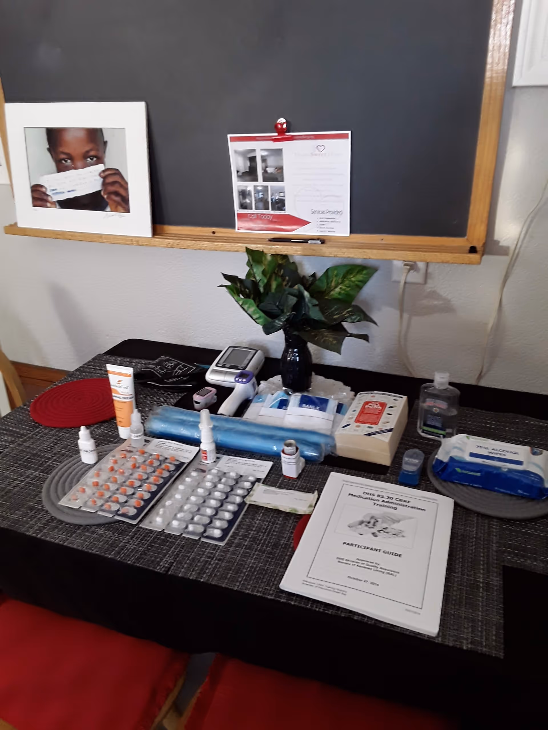 A table with various medical supplies including medication blister packs, nasal sprays, hand sanitizer, wipes, a blood pressure monitor, and a participant guide booklet for medication administration training. There is a small plant in a vase on the table and a blackboard with a photo and a flyer clipped to it in the background.