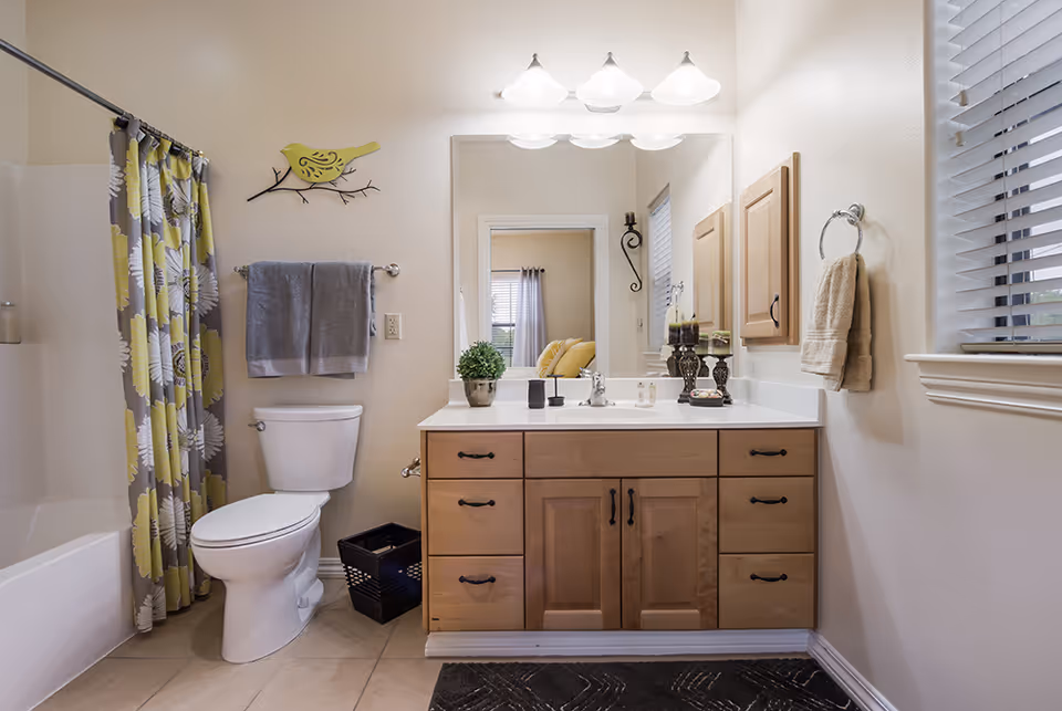 A clean and well-lit bathroom featuring a white toilet, a bathtub with a floral yellow and gray shower curtain, a wooden vanity with a white countertop, a large mirror above the sink, and decorative items including a small potted plant and candles. Towels hang on the wall, and a window with blinds is on the right side.