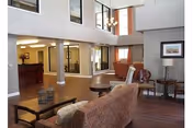 A spacious living room area with high ceilings and large windows. The room features a patterned sofa, armchairs, a coffee table, and side tables with lamps. The floor is wooden, and there is a reception desk visible in the background through a large interior window.
