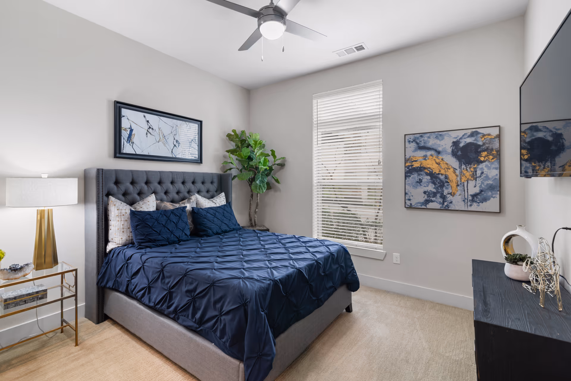 A modern bedroom with a gray upholstered bed frame and navy blue bedding. The room features a ceiling fan with a light, a window with white blinds, a bedside table with a lamp, a potted plant in the corner, abstract artwork on the walls, and a flat-screen TV mounted on the wall above a black dresser with decorative items.