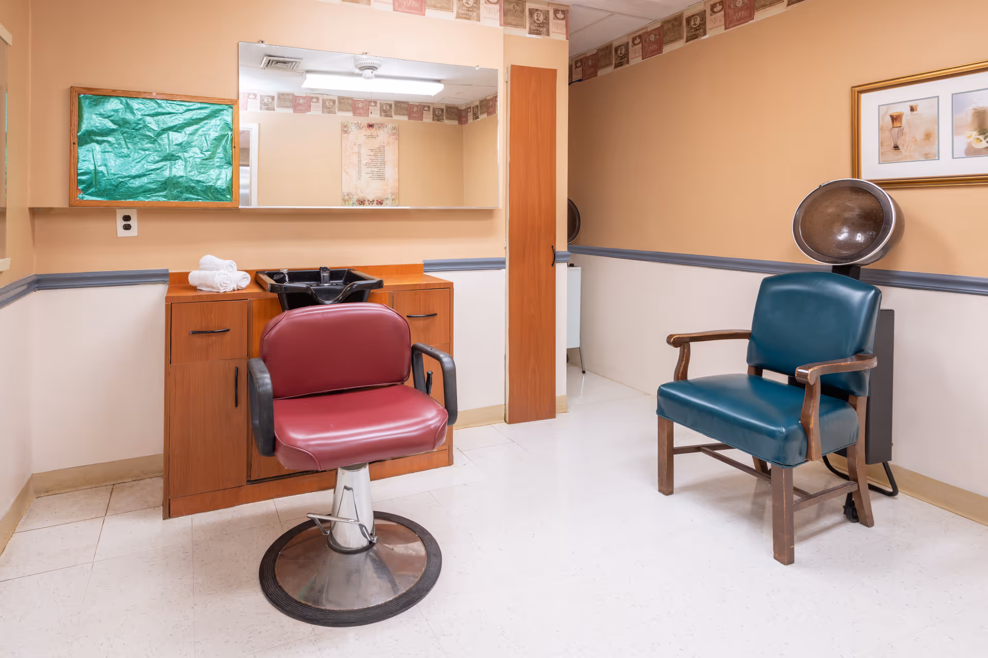 Interior view of a small salon area in a senior living facility with a maroon salon chair in front of a wooden cabinet with a black sink, a green cushioned chair with wooden armrests, a hair dryer hood, and a large mirror on the wall.